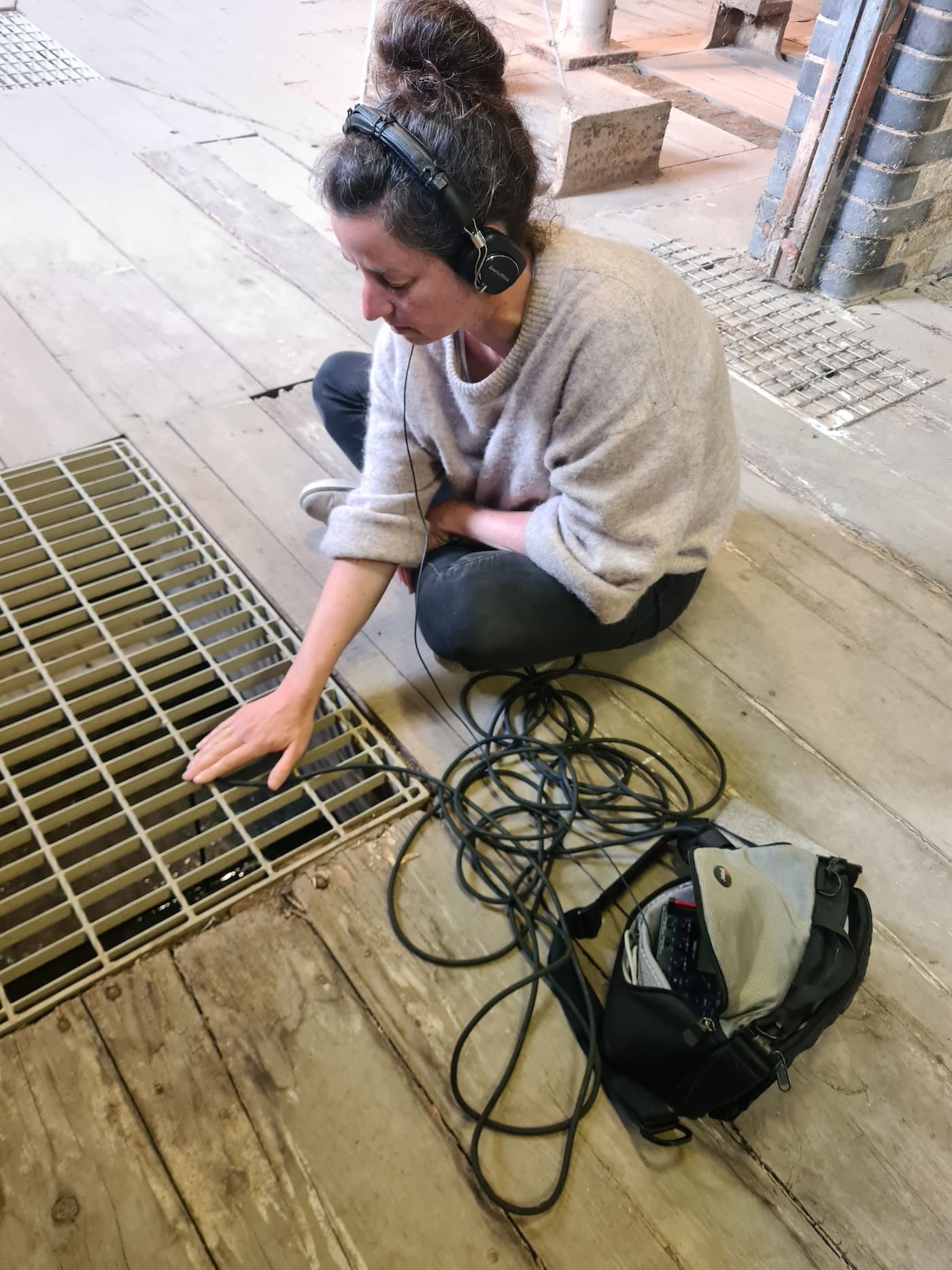 A workshop participant wearing headphones crouches on the wooden mill floor, reaching towards a metal grate to listen to the water running beneath the building