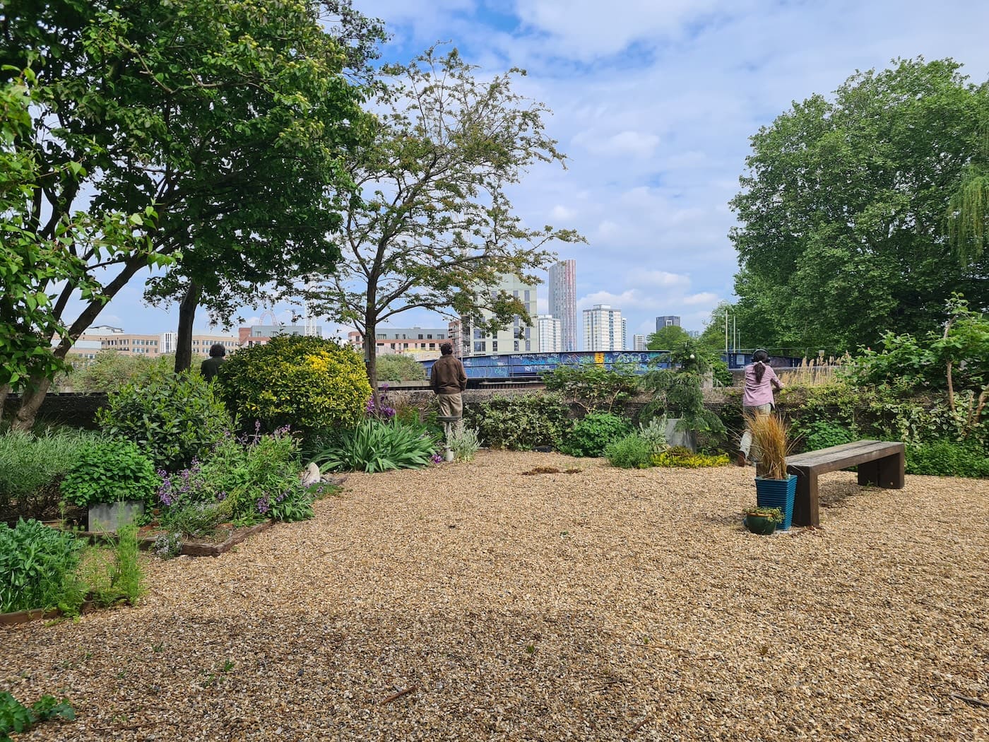 A gravel garden beside the river, where two participants look out over a hedge towards a graffiti-covered rail bridge and the Stratford skyline