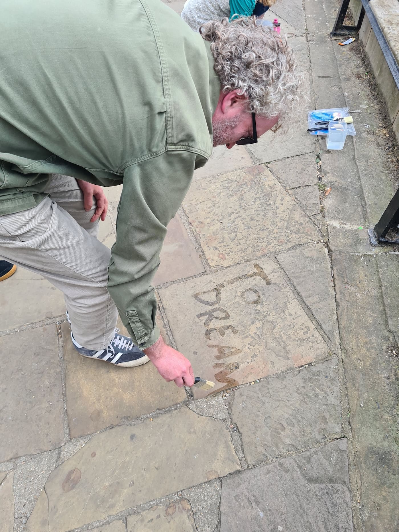 A participant bends over the pavement, brush in hand, painting the words 'To dream' onto the stone in water