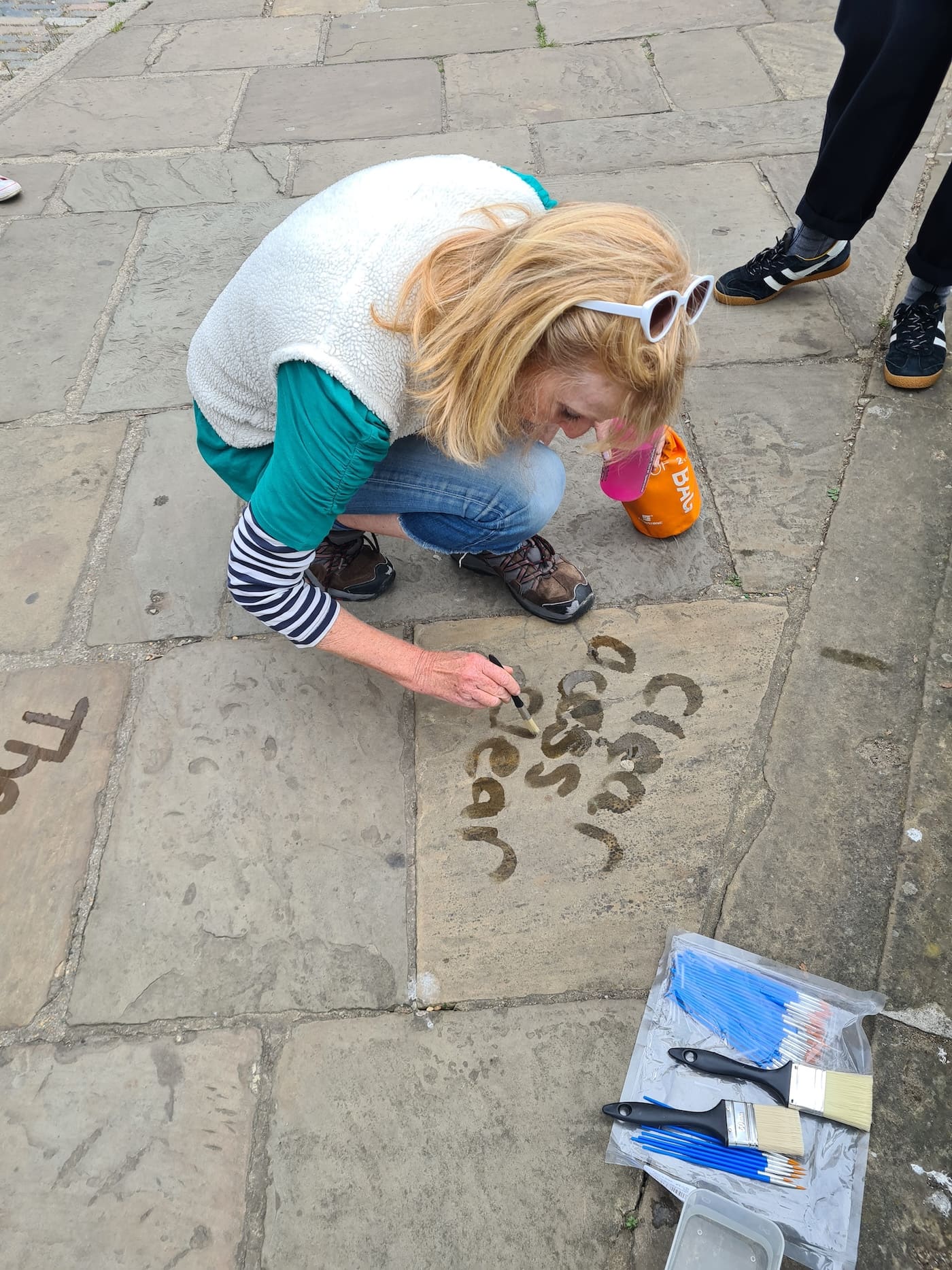 A participant crouches on the paving to write words in water with a small brush; a bag of paintbrushes lies open nearby