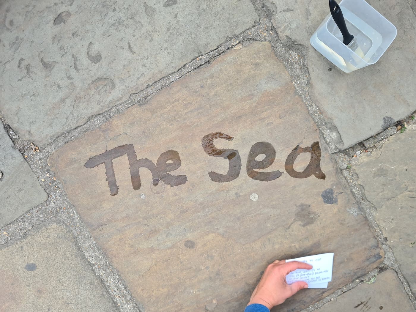 The words 'The Sea' painted in water on a paving stone, already starting to dry; a hand holds a folded slip of handwritten notes at the edge of the frame