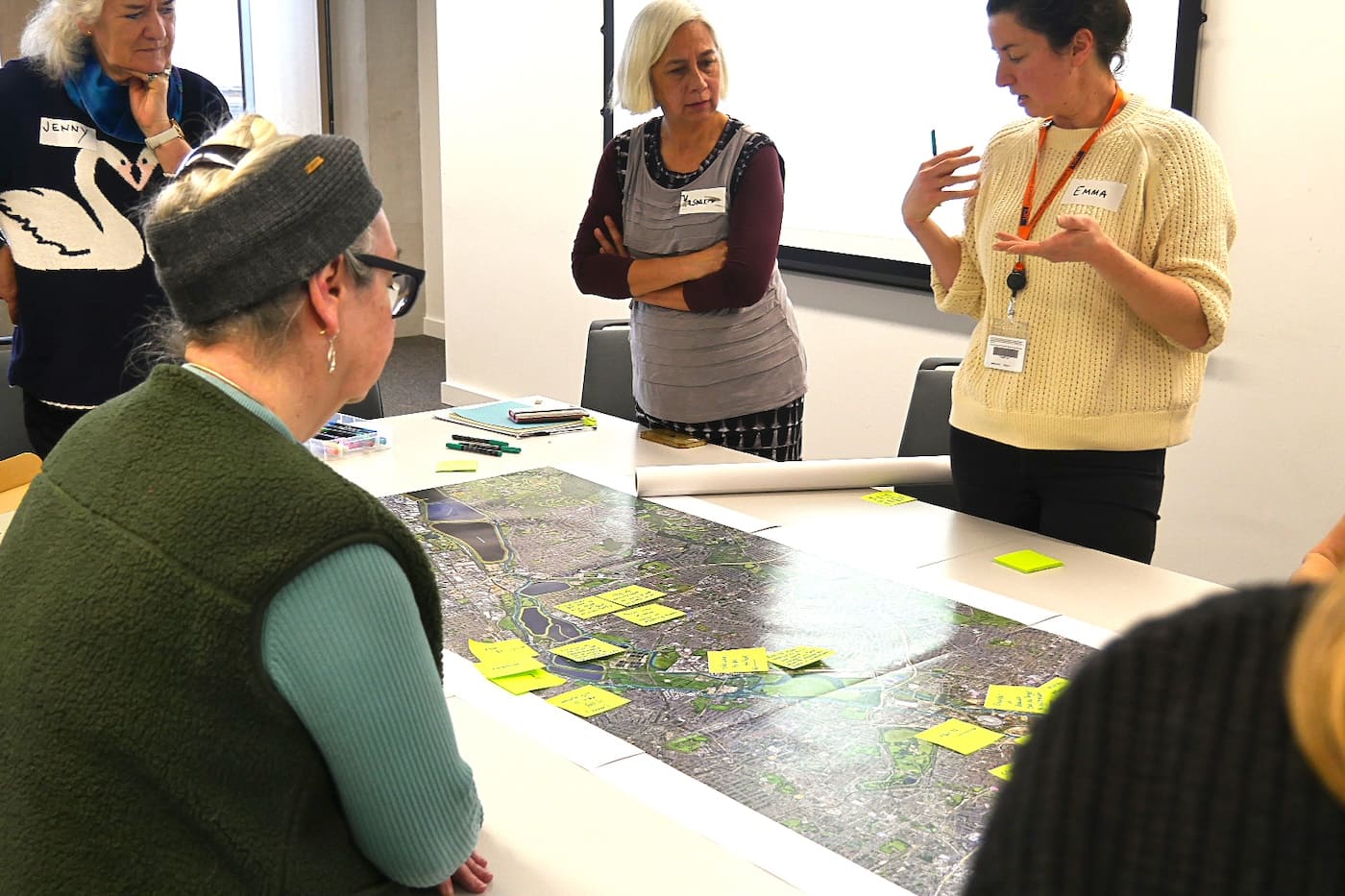 Workshop participants stand around a long aerial map of the River Lea marked with sticky notes while the artist talks it through