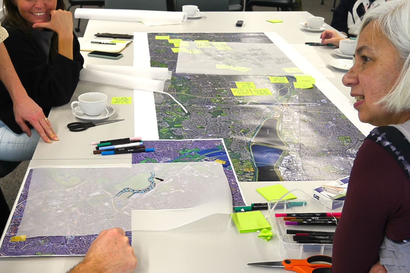 Participants around the workshop table trace the river's course onto tracing-paper overlays laid across large aerial maps