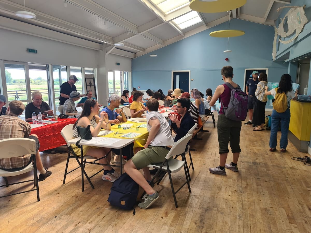 Participants seated around tables in a bright riverside community hall, talking and drawing together