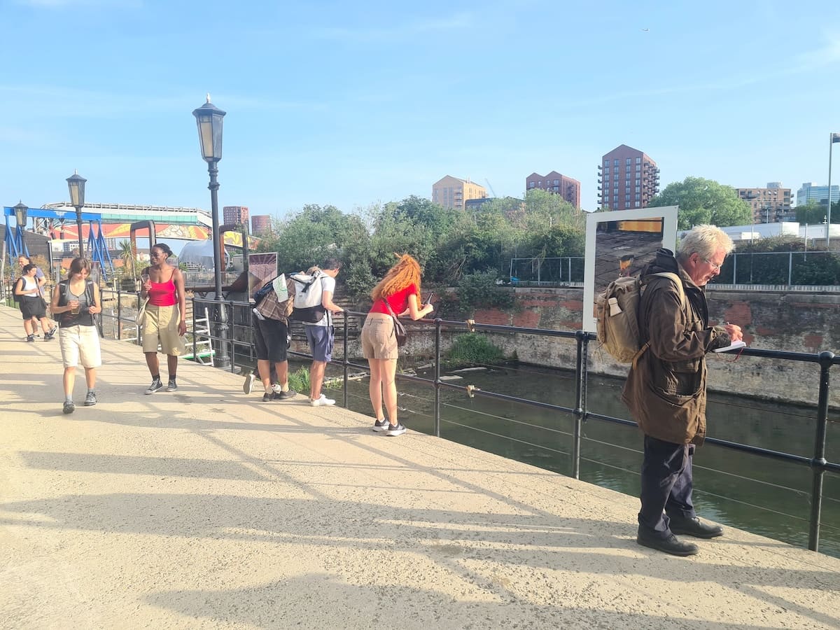 Walkers lean on the black railings of a riverside path, looking down at the water; one writes in a notebook beside a large photographic artwork mounted on the fence