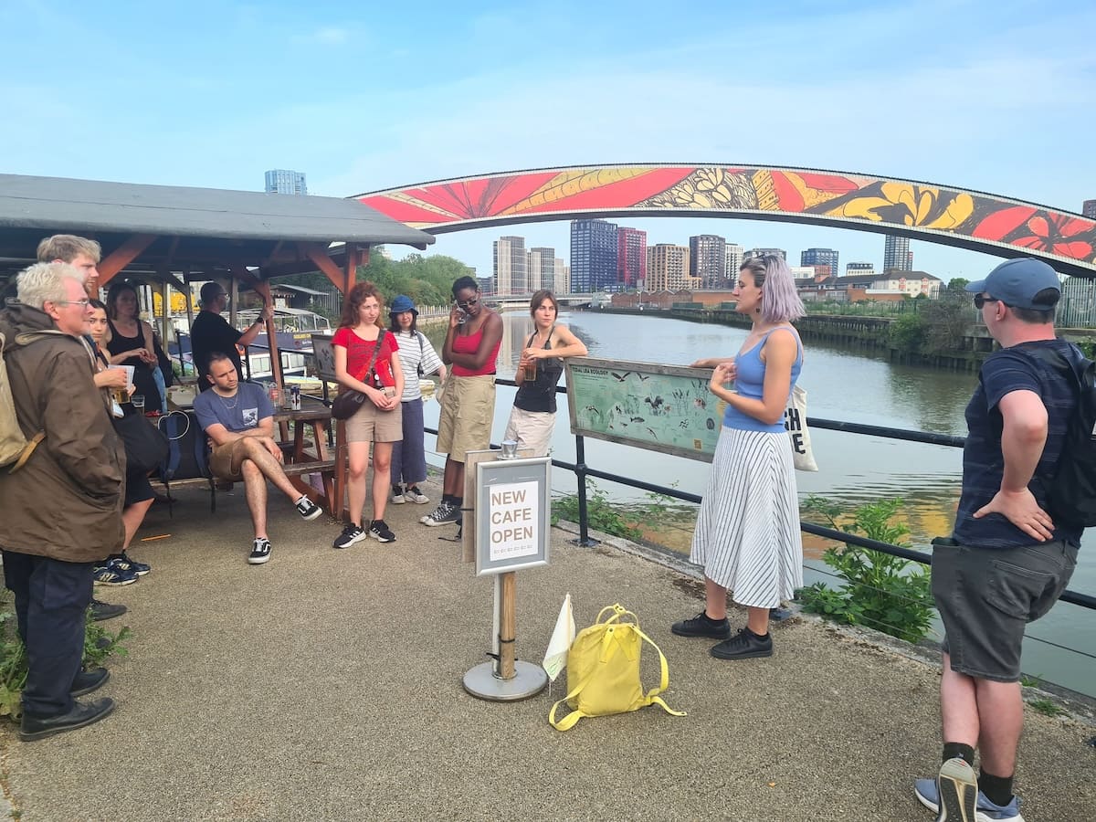 The group gathers at an ecology information board on the riverbank while a guide speaks, a butterfly-painted bridge arching over the tidal river behind