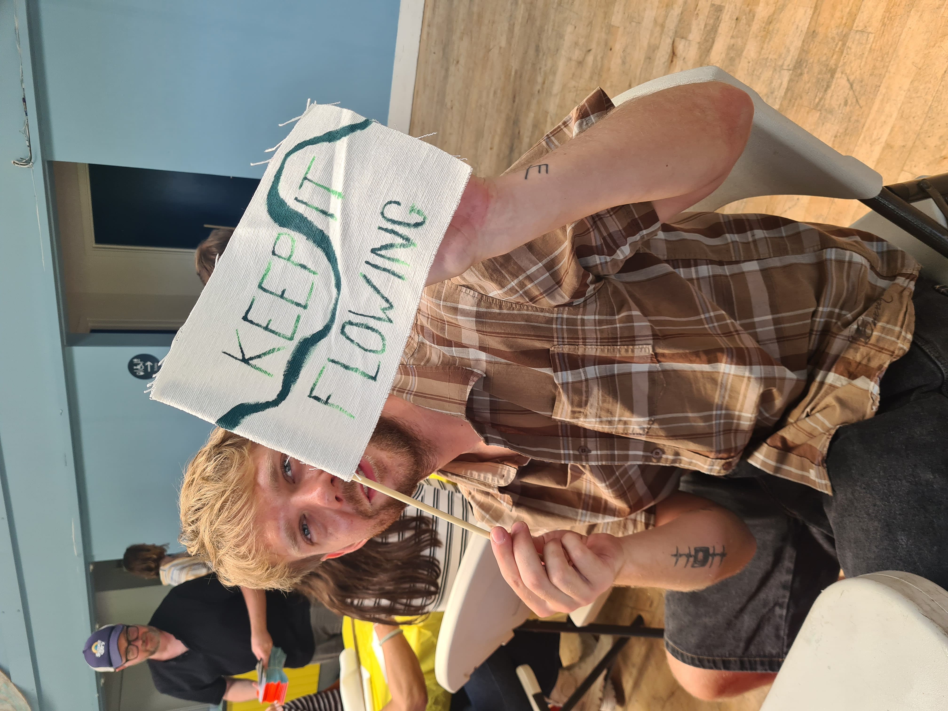 A participant holds up a small hand-painted fabric flag reading 'Keep it flowing', with a dark green river winding through the letters