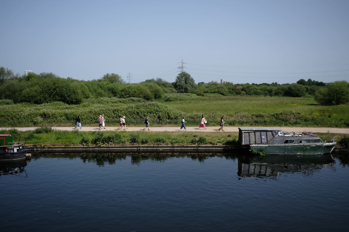 A line of walkers follows the towpath beside the River Lea past a moored boat, with open marshland stretching behind