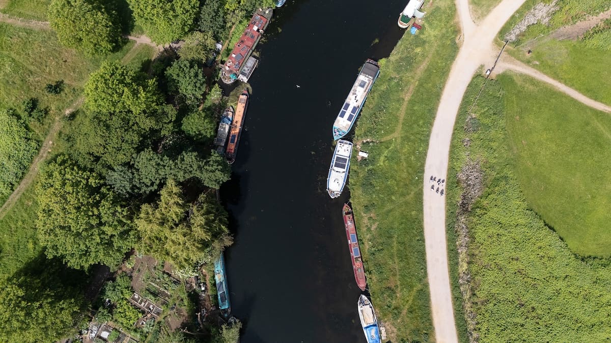 The river seen from above, narrowboats moored along both banks, as the walking group follows the pale path through the grass