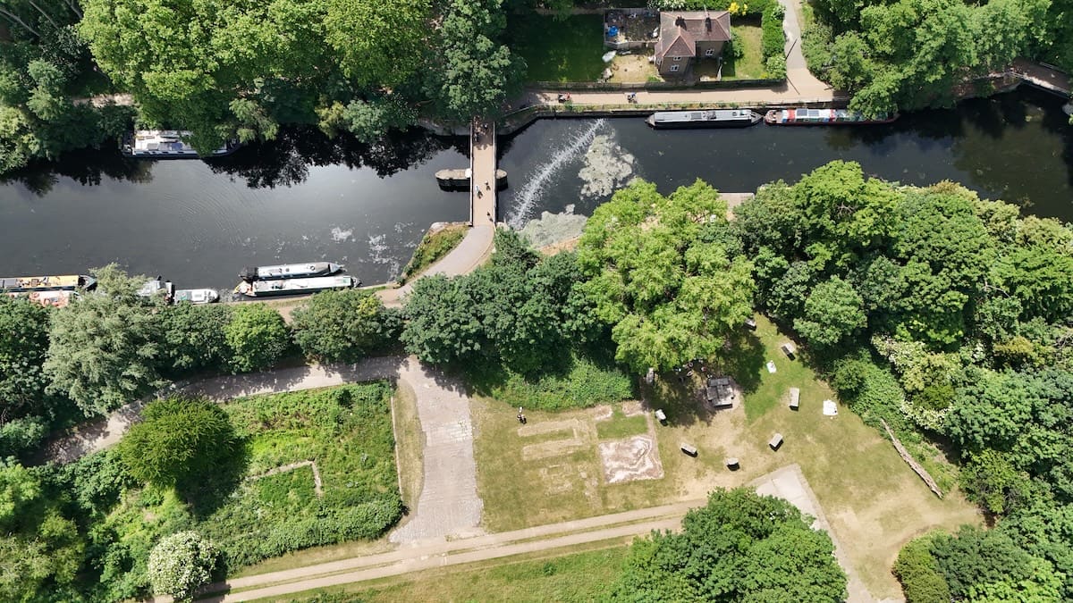 An aerial view of the river dividing at a small weir and footbridge, with narrowboats moored along tree-lined banks