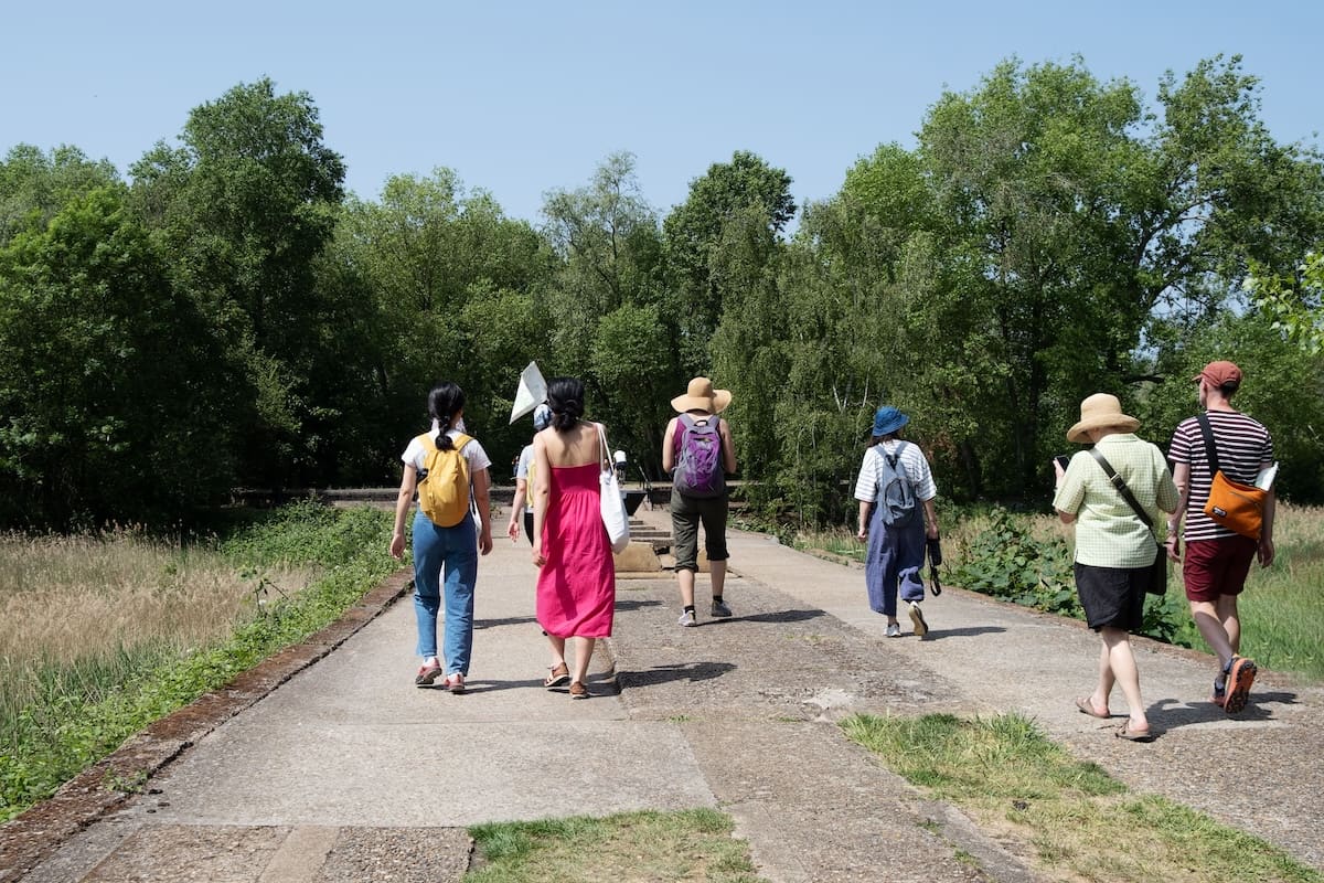 The group walks away down a broad concrete path between summer trees, sunhats on and bags over shoulders