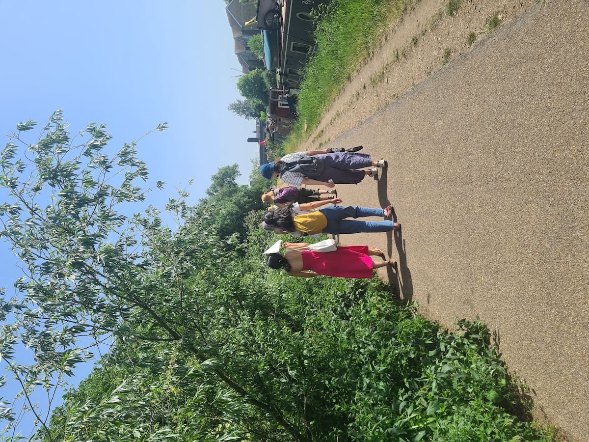 Four walkers head along a sunlit towpath beside a line of moored narrowboats, one shading herself with a folded map